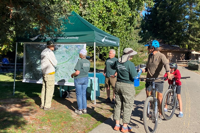 Several people and a father and son on bike stop to talk to volunteers at Water Bark event