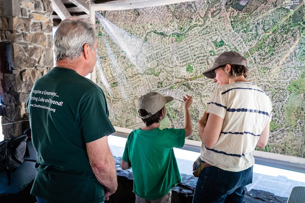 Three people, including a child in a cap pointing at a section, examine a large wall-mounted aerial map of Santa Rosa and Spring Lake County Park inside a stone pavilion.