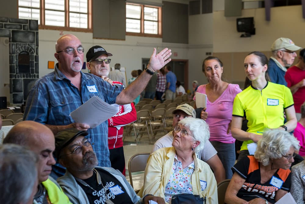 A speaker in a blue button-down shirt holds notes and raises a hand to address a crowd gathered at a community workshop.