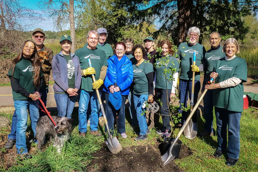 Group of Santa Rosa Southeast Greenway volunteers with shovels, young trees, and dogs outdoors on a sunny day.